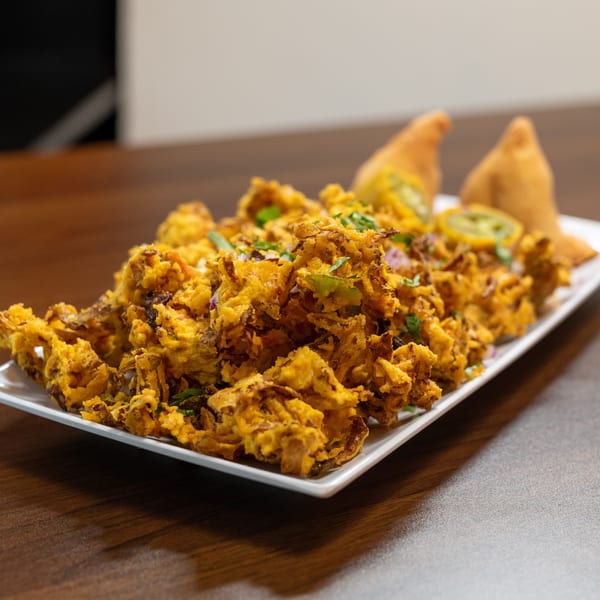 A white rectangular plate filled with golden-brown pakoras, garnished with herbs and sliced jalapeños, with two samosas in the background.