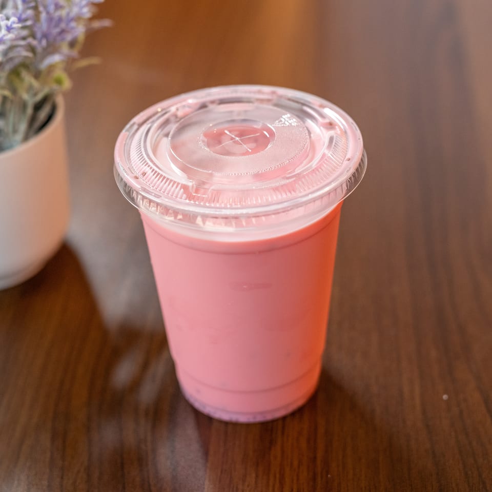 Pink beverage in a clear plastic cup with a lid, next to a potted plant on a wooden table.