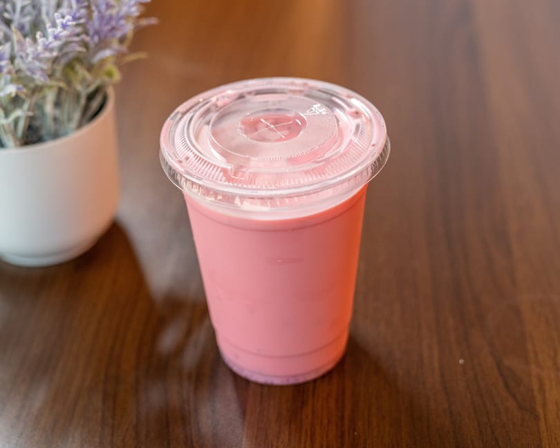 Pink beverage in a clear plastic cup with a lid, next to a potted plant on a wooden table.