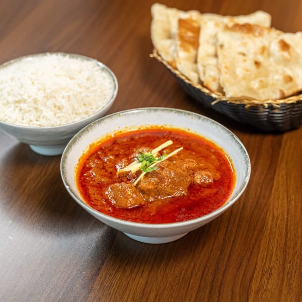 A bowl of red curry with meat, garnished with ginger and cilantro, served with rice and naan.