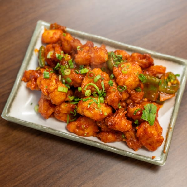 A plate of bright orange chili chicken, garnished with green onions and cilantro, on a wooden table.