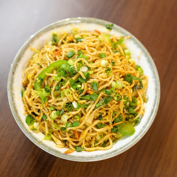 A bowl of noodles with green onions, bell peppers, and other vegetables on a wooden table.