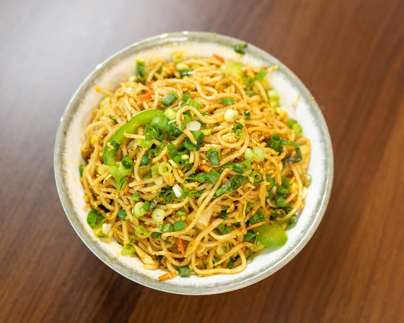 A bowl of noodles with green onions, bell peppers, and other vegetables on a wooden table.