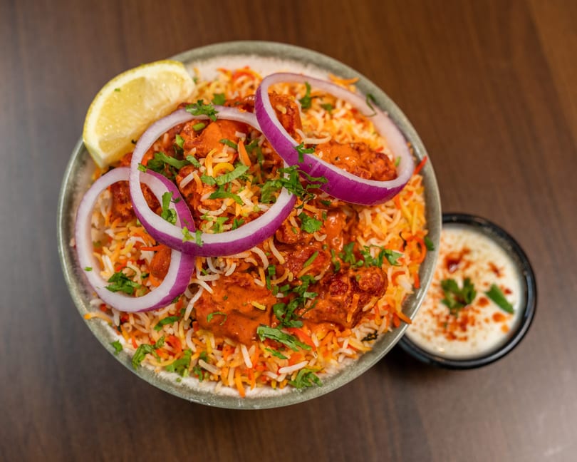 Chicken biryani with red onions, cilantro, and a lemon wedge, served with raita.