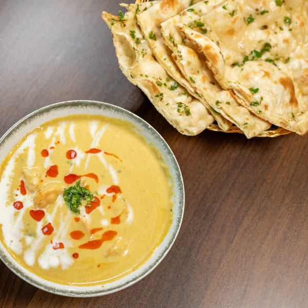 A bowl of yellow curry with white and red swirls, garnished with cilantro, next to naan bread.