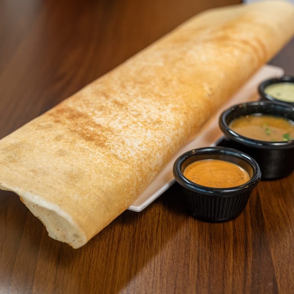 A long, golden-brown dosa on a white plate, served with three small bowls of colorful sauces.