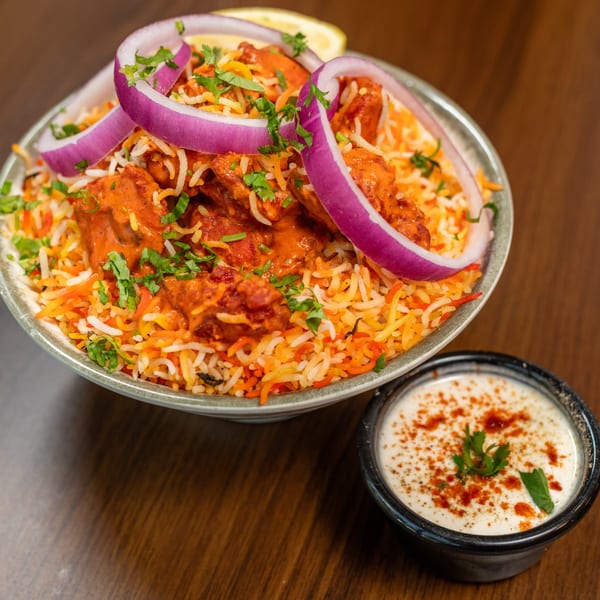 Chicken tikka biryani with red onions, cilantro, and a side of raita on a wooden table.