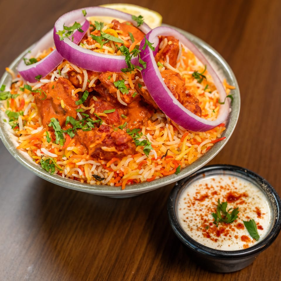 Chicken tikka biryani with red onions, cilantro, and a side of raita on a wooden table.