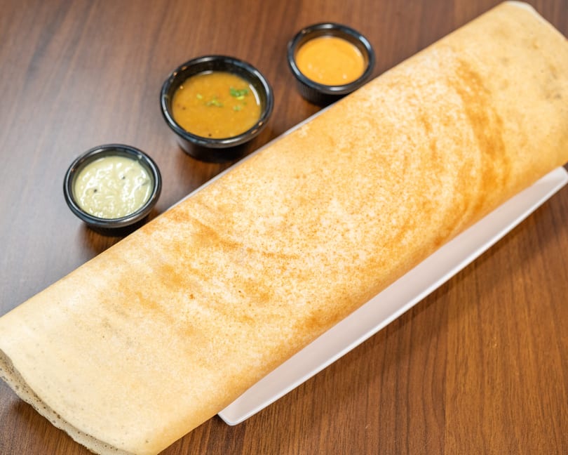 A golden-brown dosa on a white plate, served with three small bowls of chutney and sambar.
