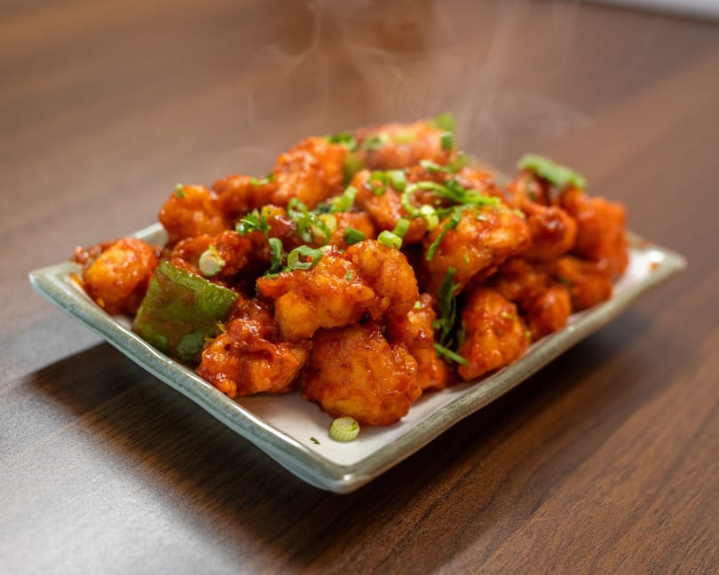 Steaming hot Gobi Manchurian, garnished with green onions and bell peppers, on a white plate.