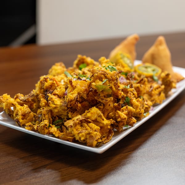 A white rectangular plate filled with golden-brown onion bhaji, garnished with herbs and two samosas.