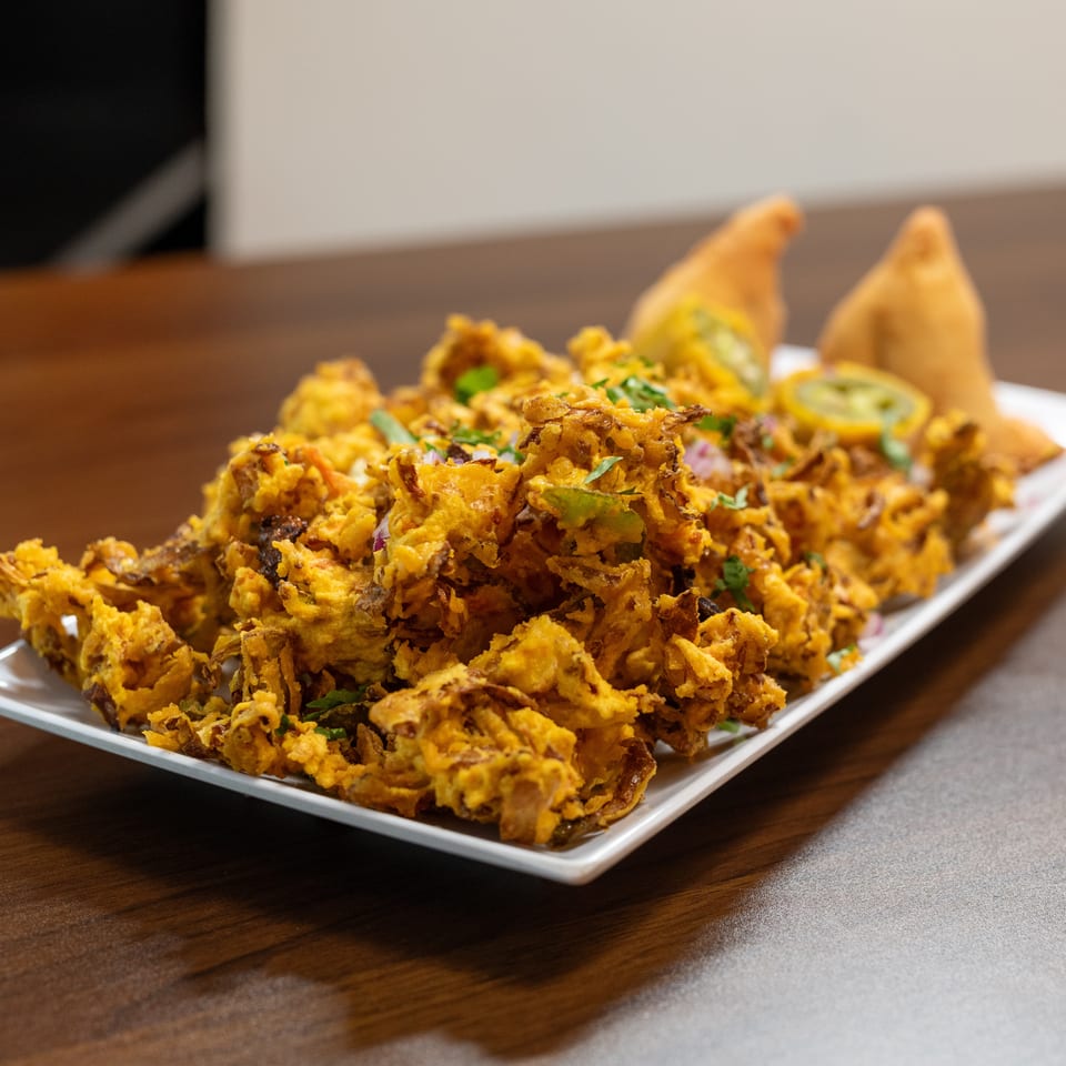 A white rectangular plate filled with golden-brown onion bhaji, garnished with herbs and two samosas.