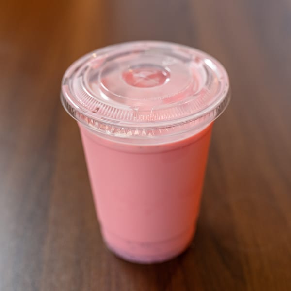 Pink strawberry milkshake in a clear plastic cup with a lid on a wooden table.