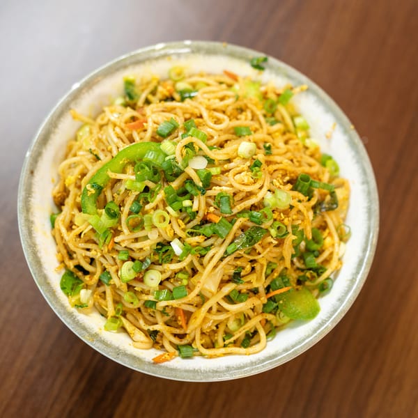 A bowl of noodles with green onions, bell peppers, and other vegetables on a wooden table.