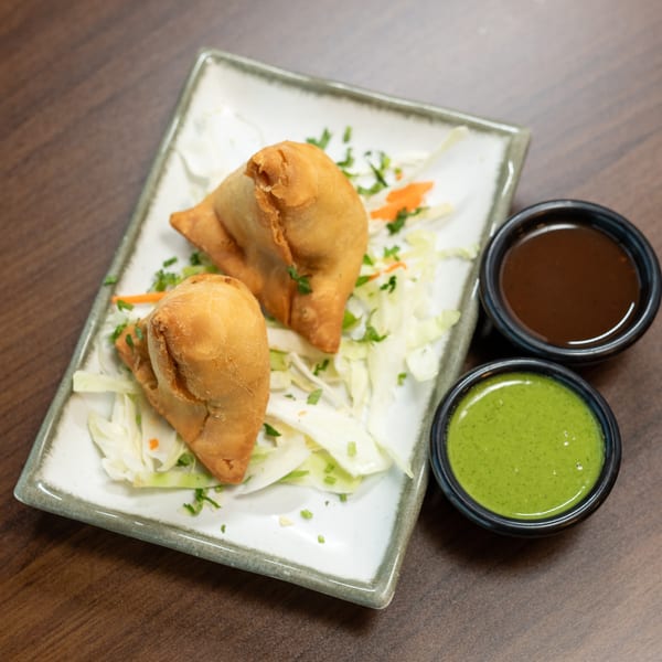 Two golden-brown samosas on a bed of shredded cabbage, with green and brown dipping sauces.