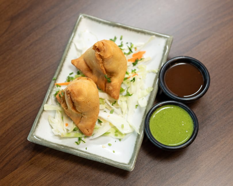 Two golden-brown samosas on a bed of shredded cabbage with green and brown dipping sauces.